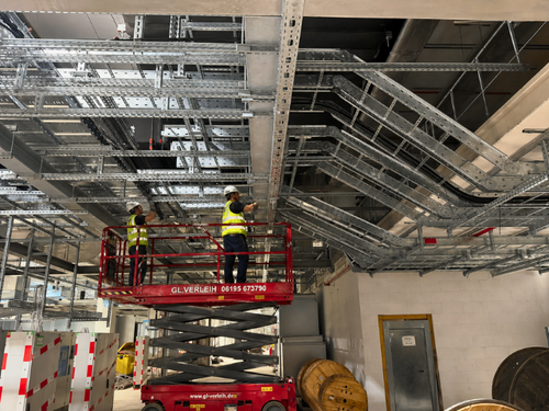 Instaladores trabajando en altura sobre plataformas elevadoras, revisando cableado eléctrico sobre bandejas portacables metálicas PohlCon en el techo técnico de una nave industrial.